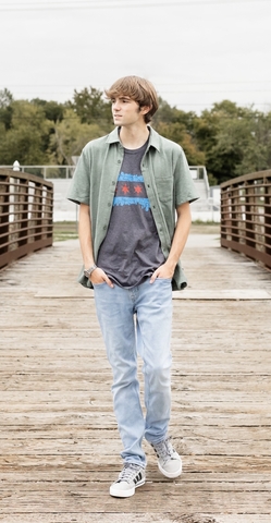 student posed on a bridge with a gray shirt and green button up shirt and blue jeans.