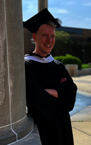 Christopher in his master's cap and gown leaning against a pillar.