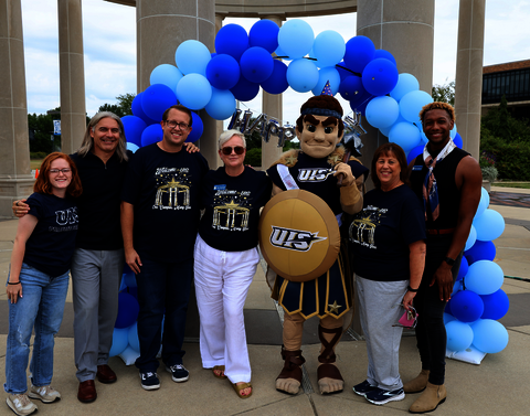A group of people standing with a mascot under a balloon arch.