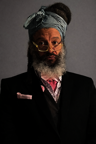 Man with beard and turban, wearing glasses and a black suit with a pink tie.