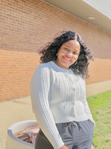 student posed in front of a brick wall with a gray sweater and dark gray pants.