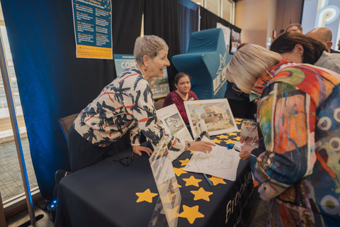 A woman at a table hands papers to another woman, surrounded by star shapes.