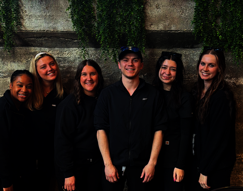 Six people pose in all black in front of a stone wall.