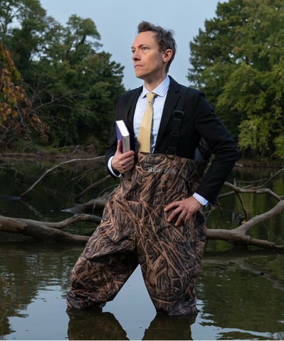 University Scholar Brandon Derman standing in Lake Springfield in waders and a suit and holding a book.