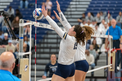 Two UIS volleyball players in mid-air blocking a ball at the net