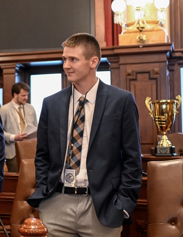 student posed on the state capitol house/senate floor with a Navy blue suit jacket, white button down and tie.