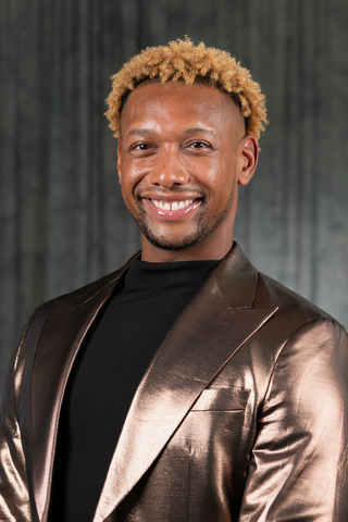Smiling person in a shiny bronze blazer against a dark background.