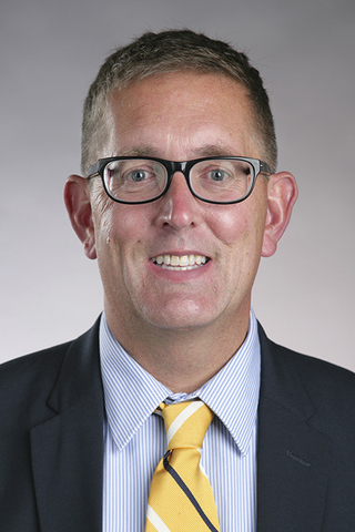 Man wearing glasses, a suit, and a yellow striped tie, smiling against a gray background.