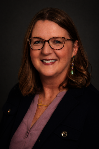 Professional headshot of Tami Ireland smiling against a gray background, wearing glasses, a navy blazer, and a pink blouse.
