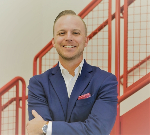 Professional headshot of Joshua Frye smiling with his arms crossed, wearing a blue suit jacket and white shirt in front of a red staircase railing.