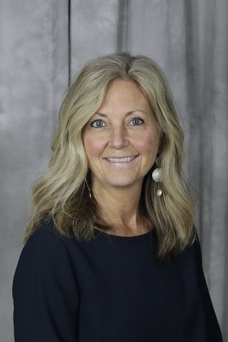 Professional headshot of Susan O’Malley smiling against a gray patterned background, wearing a navy blue top and dangling earrings.