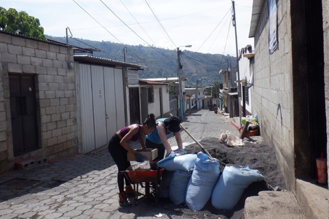 Two students sift sand into a wheelbarrow on a narrow village street lined with homes in Guatemala.