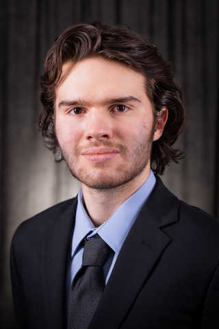 Professional headshot of Joe wearing a blue button-down shirt, black blazer, and dark gray tie with a neutral background.