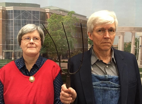 A woman standing next to a man dressed in overalls who is holding a pitch fork.