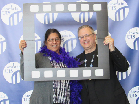 A man and a woman standing in front of a backdrop that reads "I Invest in UIS" holding a film strip cut out. The woman is wearing a purple feather boa.