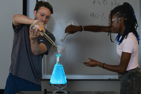 Students conduct a chemistry experiment, pouring liquids into a beaker with glowing blue liquid under safety precautions.