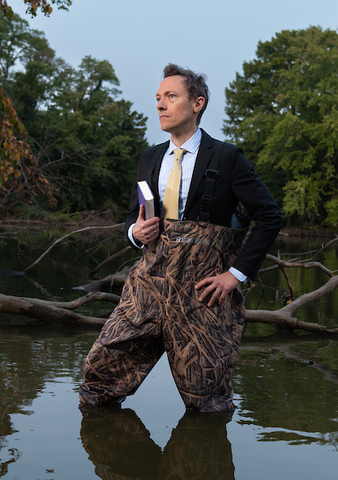 Associate Professor Brandon Derman wears a suit coat and tie with camouflaged fishing waders and stands in a placid pool of green water with trees and limbs in the background.