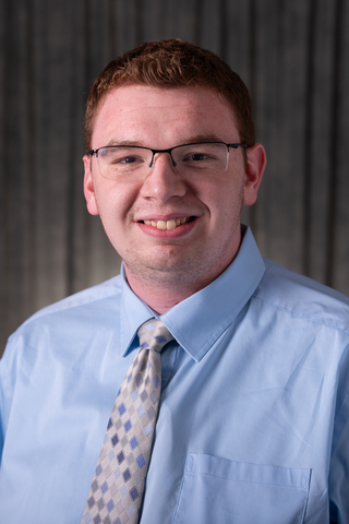 Professional headshot of Caleb wearing a blue button-down shirt and gray and blue tie, smiling with a neutral background.