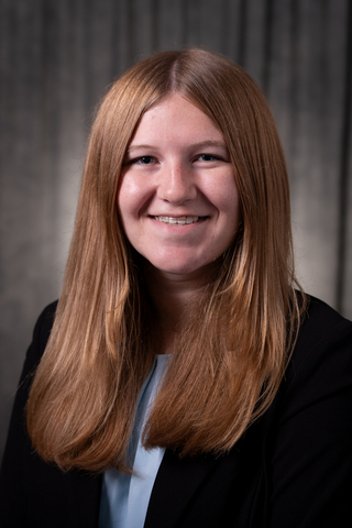 Professional headshot of Corinna wearing a light blue shirt and black blazer, smiling with a neutral background.