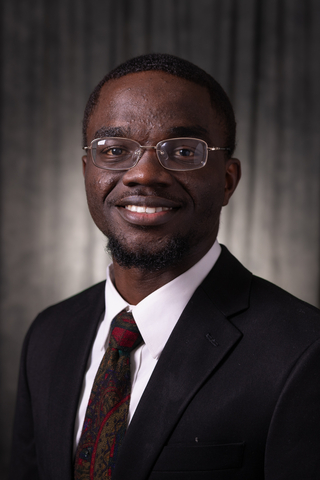 Professional headshot of Sly wearing a white button-down shirt, black blazer, and red tie with designs smiling with a neutral background.