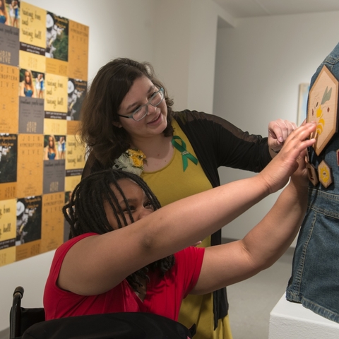 A female in a wheelchair feels a design in wood on the back of a jeans jacket in an art exhibition while another female describes what she is feeling.