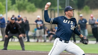 Quinn Gudaitis pitches in the foregrown in a dark blue UIS jersey while a line judges stands by in the background.