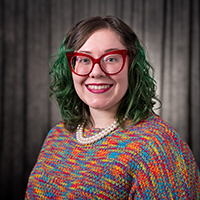 Dre Duvendack Visk, a smiling woman with glasses and colorful sweater against a dark background.