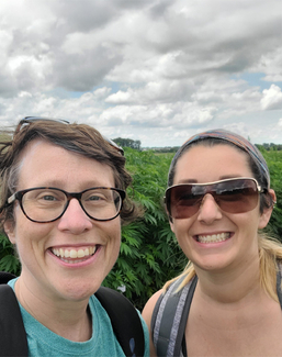 Two people smiling outdoors under a cloudy sky, surrounded by greenery.