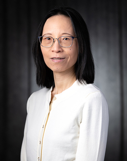 Woman with glasses and a white cardigan, dark background, neutral expression.