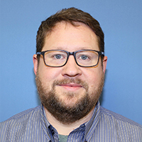 Jeff Sudduth, a man with short brown hair, beard, and glasses wearing a striped shirt, photographed against a plain blue background.