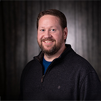 Jeff Sudduth, a smiling man in dark sweater against a dark background.