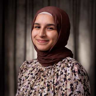 Smiling person in a floral blouse and dark hijab in front of a grey background.