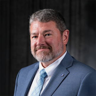 Man with a beard in a blue suit and tie against a dark background.
