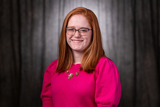 Smiling person with red hair and glasses, wearing a pink sweater, against a dark backdrop.