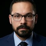 Man with glasses and beard wearing a suit in dark lighting.