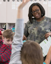 A teacher in a classroom smiling while students raise their hands.