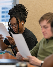 Two students sitting at a desk in a classroom reading pieces of paper. 