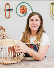 A woman with long hair works on weaving a wicker basket at a wooden table. Behind her, colorful, abstract wall art creates a creative atmosphere.