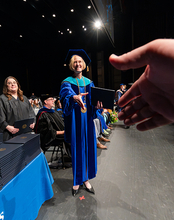 A university official in academic regalia extends a diploma and handshake to a graduate during a commencement ceremony.