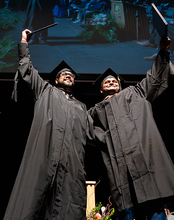 Two graduates in black caps and gowns celebrate on stage by raising their diplomas triumphantly, smiling as they stand side by side. Behind them, faculty members in academic regalia sit in rows and a large screen displays a projection of the moment above them.