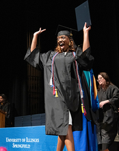 Graduating student celebrates on stage in cap and gown holding diploma at UIS Commencement as faculty applaud.
