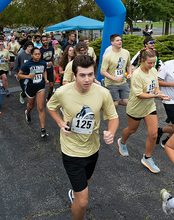 A group of runners participates in a race, starting under a blue inflatable arch. Participants wear numbered bibs and light-colored shirts.