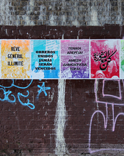 A vibrant poster series on a graffiti-covered brick wall. Each colorful panel displays multilingual text.