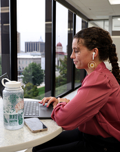 Young woman works on a laptop at a high-rise office window with earbuds, water bottle, and phone nearby.