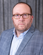 Man in glasses wearing a blue blazer and shirt, against a dark curtain backdrop.