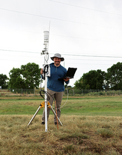 Man with tablet stands by weather station in grassy field.