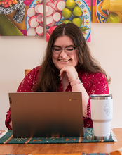 Woman smiling at a laptop with a large mug on the table; colorful art in the background.