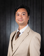 Man in a tan suit with a tie, black background, smiling gently.