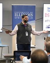 Man speaking at a presentation, University of Illinois Springfield banner behind him.
