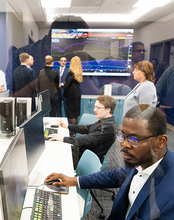 People working at computers in an office with a stock market screen.